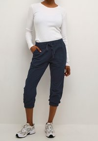 Pantalon de jogger court bleu marine avec taille réglable à cordon, associé à un t-shirt blanc ajusté à manches longues et des chaussures de sport de couleur claire.