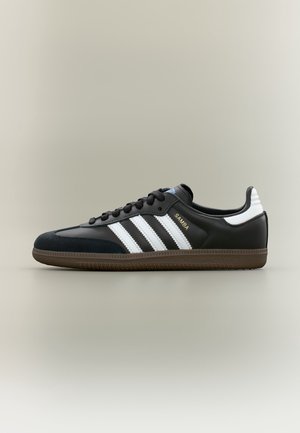 Baskets Adidas Samba noires avec des bandes blanches, un logo doré, une semelle en gomme et des lacets noirs, vues de profil sur un fond clair.