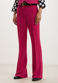 Pantaloni su misura fucsia realizzati in tessuto liscio, caratterizzati da un taglio a gamba larga e dettagli di pieghe, abbinati a scarpe nere.