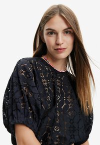 Sort blonde top med et blomstermønster og puffede ærmer. Sammen med en farverig perlekæde, der fremhæver det intrikate design og tekstur.