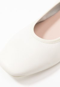 Vita läderskor med platt sula, rundad fyrkantig tå och elastisk öppning. Slät yta med en mjuk, ljusrosa innersula.