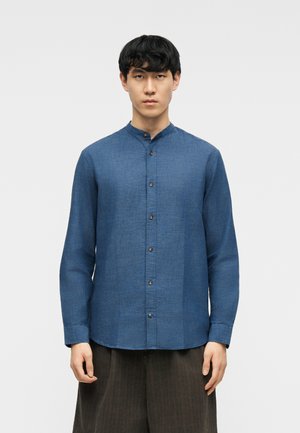 Uomo che indossa una camicia blu con colletto alla coreana abbottonata e maniche lunghe, e pantaloni larghi scuri, in piedi contro uno sfondo chiaro e semplice.