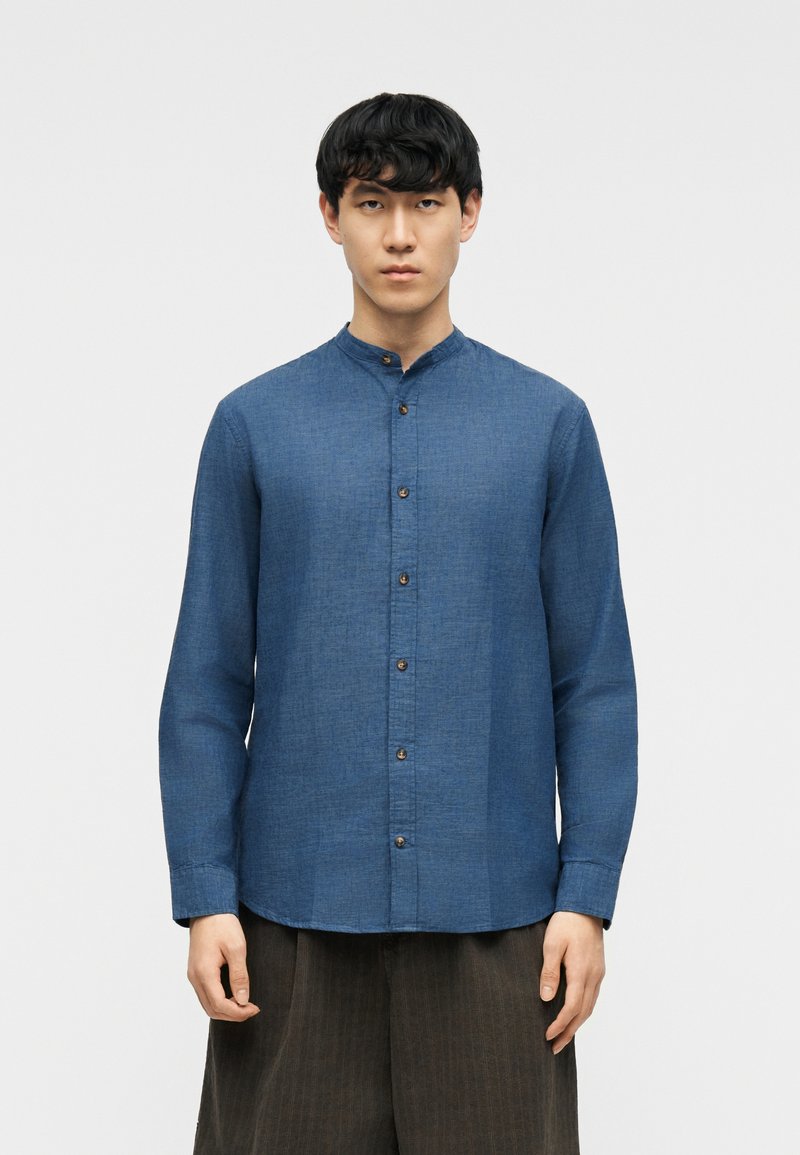 Άνδρας που φοράει μπλε πουκάμισο με όρθιο γιακά και κουμπιά, μακριά μανίκια και σκούρο παντελόνι με φαρδιά πόδια, στέκεται μπροστά σε ένα απλό φωτεινό φόντο.