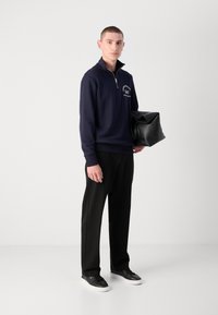Navy zip-up sweatshirt with white graphic text, paired with black trousers and black sneakers. Model holds a black bag.