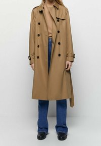 Beige trench coat made of lightweight fabric with a double-breasted design, black buttons, and a tie belt, paired with blue flared jeans.