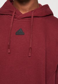 Maroon huppari, jossa on naru ja pieni musta Adidas-logo patch rintamuksessa. Valmistettu pehmeistä kankaista ja rento muotoilu.