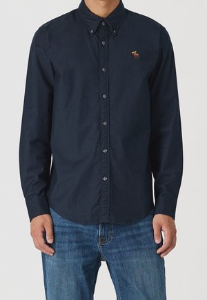 Mand iført en mørk marineblå skjorte med knapper og en lille elg-logo på brystet samt blå jeans, stående med armene afslappet ned langs siderne.