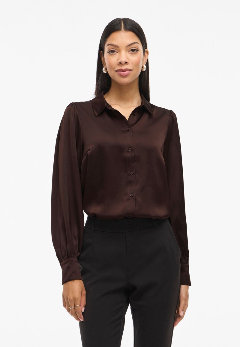 Smeđa satenska bluzica s ovratnikom, prednjim gumbima i dugim rukavima. Glatka tekstura s laganim sjajem, nosi se uz crne krojene hlače.