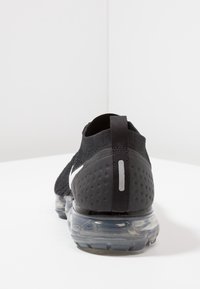 Chaussure de sport noire en matériau tricoté avec un talon à motif, une unité d'air transparente et un logo argenté réfléchissant à l'arrière.