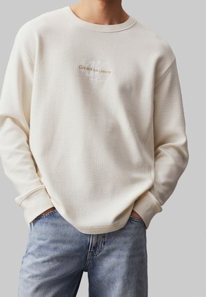 Mężczyzna noszący kremową, teksturowaną koszulę Calvin Klein Jeans z długim rękawem, ręce w kieszeniach oraz jasnoniebieskie jeansy na jednolitym tle.