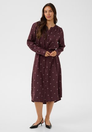 Femme aux longs cheveux bruns portant une robe à manches longues, boutonnée, de couleur violet foncé avec de petits motifs et des chaussures noires à talons bas.