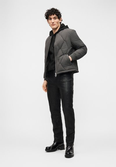Νέος άνδρας που στέκεται ντυμένος με μαύρο δερμάτινο παντελόνι, μαύρο φούτερ με κουκούλα, γκρι καπιτονέ μπουφάν και μαύρα λαμπερά μπότες σε λευκό φόντο.