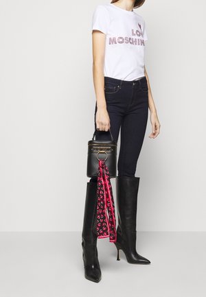 Czarna torebka na ramię z uchwytem i złotymi elementami, z dołączonym czerwono-czarnym wzorzystym szalem. Modelka ma na sobie białą koszulkę i ciemne dżinsy.