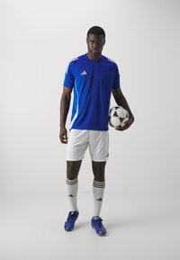 Blå treningstrøye med hvite detaljer, hvite shorts, blå fotballsko og strømpene til knærne med striper; holder en svart og hvit fotball.