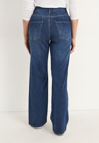 Kvinna som bär högmidjade, vida mörkbl�å jeans och en vit långärmad topp, sedd bakifrån mot en enfärgad bakgrund.