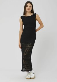 Robe longue noire en crochet avec un corsage ajusté, des épaules larges et un bas transparent. Associée à des baskets blanches. Texture lisse et tissage ouvert.