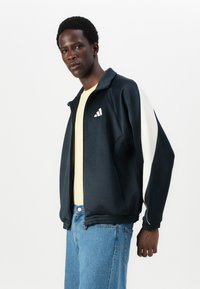 Casaco desportivo em azul-marinho e creme com colarinho alto e fecho éclair completo. Apresenta um logótipo branco e punhos canelados. Usado sobre uma camisa amarela e jeans azuis.