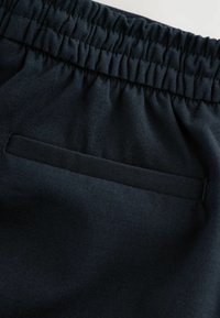Tmavě modrá látka s hladkým povrchem. Obsahuje elastický pas a klopovou kapsu, což spojuje funkčnost s minimalistickým designem.