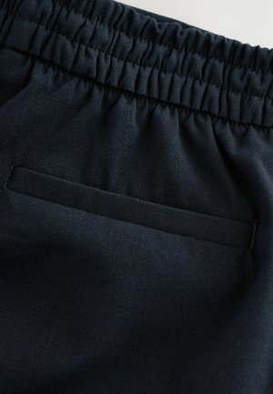 Tmavě modrá látka s hladkým povrchem. Obsahuje elastický pas a klopovou kapsu, což spojuje funkčnost s minimalistickým designem.