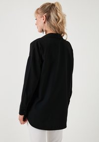 Cămașă neagră cu mâneci lungi, cu o croială lejeră, cu manșete buton închise, și un guler clasic, având o textură fină și un tiv rotunjit.