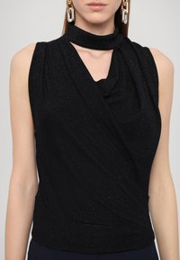 Haut noir sans manches en tissu scintillant, encolure drapée et col étroit semblable à une écharpe, porté par une personne avec de grandes boucles d'oreilles métalliques.