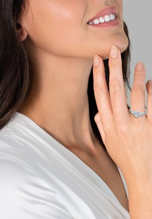 Silberner Verlobungsring mit einem zentralen Edelstein, filigranen Ringdetails, getragen an einer Hand mit heller Haut, vor einem neutralen Hintergrund.