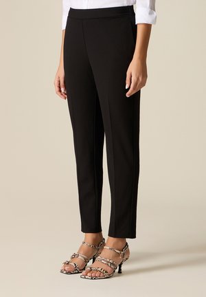 Kvinde iført skræddersyede sorte ankellange bukser med hælesandaler med slange-mønster og en hvid skjorte med opkærmede ærmer.