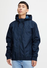 Navy blue huppari, joka on valmistettu vedenpitävästä materiaalista, varustettuna vetoketjullisella rintataskulla, elastisilla hihansuilla ja narupäällä.