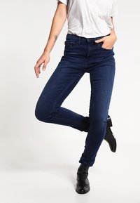 Jean skinny bleu marine avec une coupe ajustée, design à cinq poches et taille mi-haute, associé à des bottines noires.