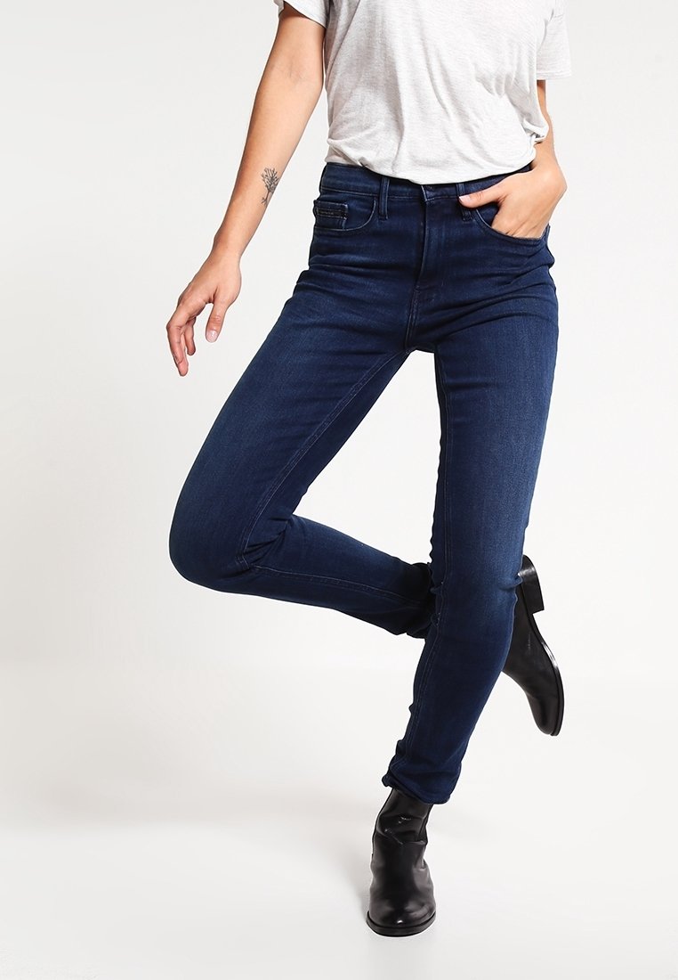 Jean skinny bleu marine avec une coupe ajustée, design à cinq poches et taille mi-haute, associé à des bottines noires.