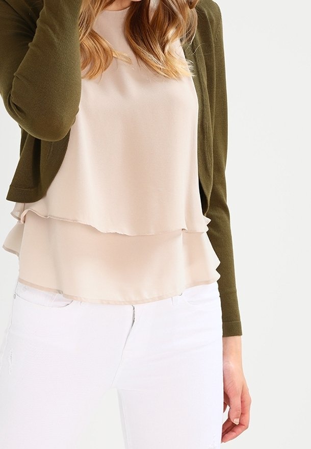 Femme portant un cardigan vert olive par-dessus un chemisier beige superposé et un pantalon blanc sur un fond uni.