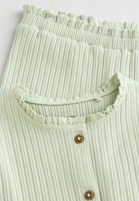 Grünes geripptes Oberteil mit Rüschenausschnitt und zwei holzähnlichen Knöpfen, kombiniert mit passenden gerippten Hosen mit hoher Taille. Weicher Stoff, leichtes Material.