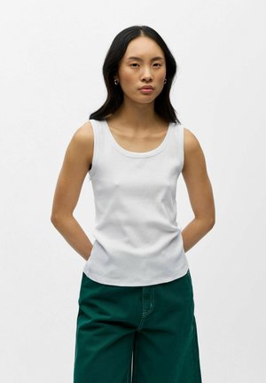 Jeune femme aux longs cheveux noirs portant un haut blanc sans manches et un pantalon vert, debout les mains derrière le dos sur un fond blanc.