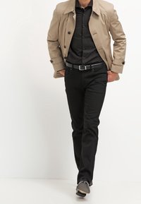 En tanfärgad trenchcoat över en svart knapp-up skjorta, kombinerat med svarta byxor och grå skor. Bältet har en silverfärgad spänne.