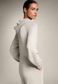 Felpa grigia chiara con fit rilassato, presenta un cappuccio con cordoncino, maniche lunghe e tasche laterali. Materiale morbido e elastico.