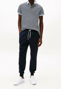 Polo à manches courtes, rayé bleu marine et blanc avec un col bleu clair, associé à un pantalon de jogging noir avec cordon de serrage et chevilles resserrées, ainsi que des baskets blanches.