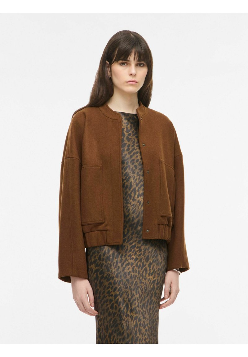 Femme portant une veste courte marron sur une robe marron et noire à imprimé léopard, debout devant un fond blanc uni.