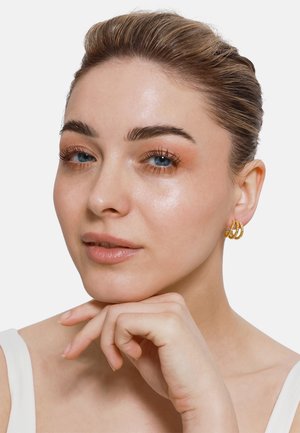 Goldene Ohringe mit drei strukturierten Ringen, die eine glänzende Oberfläche aufweisen. Das Model hat kurze Haare und trägt ein weißes Oberteil. Die Haut wirkt strahlend.