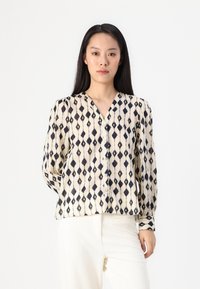 Cream blouse with navy diamond pattern, V-neck, buttons down the front, and long puff sleeves. Worn with light-colored textured pants.
