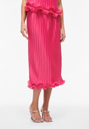 Kobieta nosząca jasnoróżową plisowaną spódnicę midi z falbaną na dole oraz srebrne sandały na wysokim obcasie z odkrytymi palcami na białym tle.