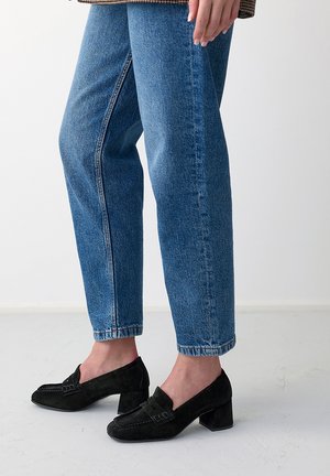 Personne portant un jean court bleu et des mocassins noirs en daim à talon bloc, debout sur un sol clair devant un fond blanc uni.