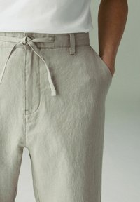 Pantaloni di lino grigio chiaro con vestibilità comoda, vita con cordoncino e tasche laterali. La texture sembra morbida e leggermente stropicciata.