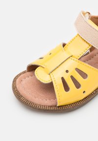 Sandal in vernice gialla con cinturino in suede beige, suola interna morbida e dettagli decorativi a intaglio sul cinturino anteriore.