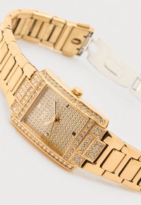 Rechthoekig horloge met goudkleurige afwerking, voorzien van een met kristallen bezette wijzerplaat en lunette. De metalen schakelarmband heeft een gepolijste afwerking en een veilig sluitsysteem.