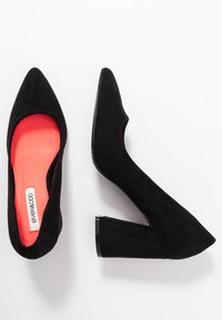 Black suede high heels with a pointed toe and a wide block heel. Interior features a bright coral insole. Smooth texture, minimalist design.
