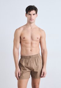 Boxeri din bumbac maro, cu talie elastică, având o croială lejeră și o textură netedă. Purtați de un model cu o constituție atletică.