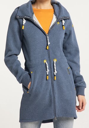 Femme portant un hoodie bleu long à fermeture éclair avec cordons blancs et accents jaunes, sur un t-shirt orange et un jean, les mains dans les poches.