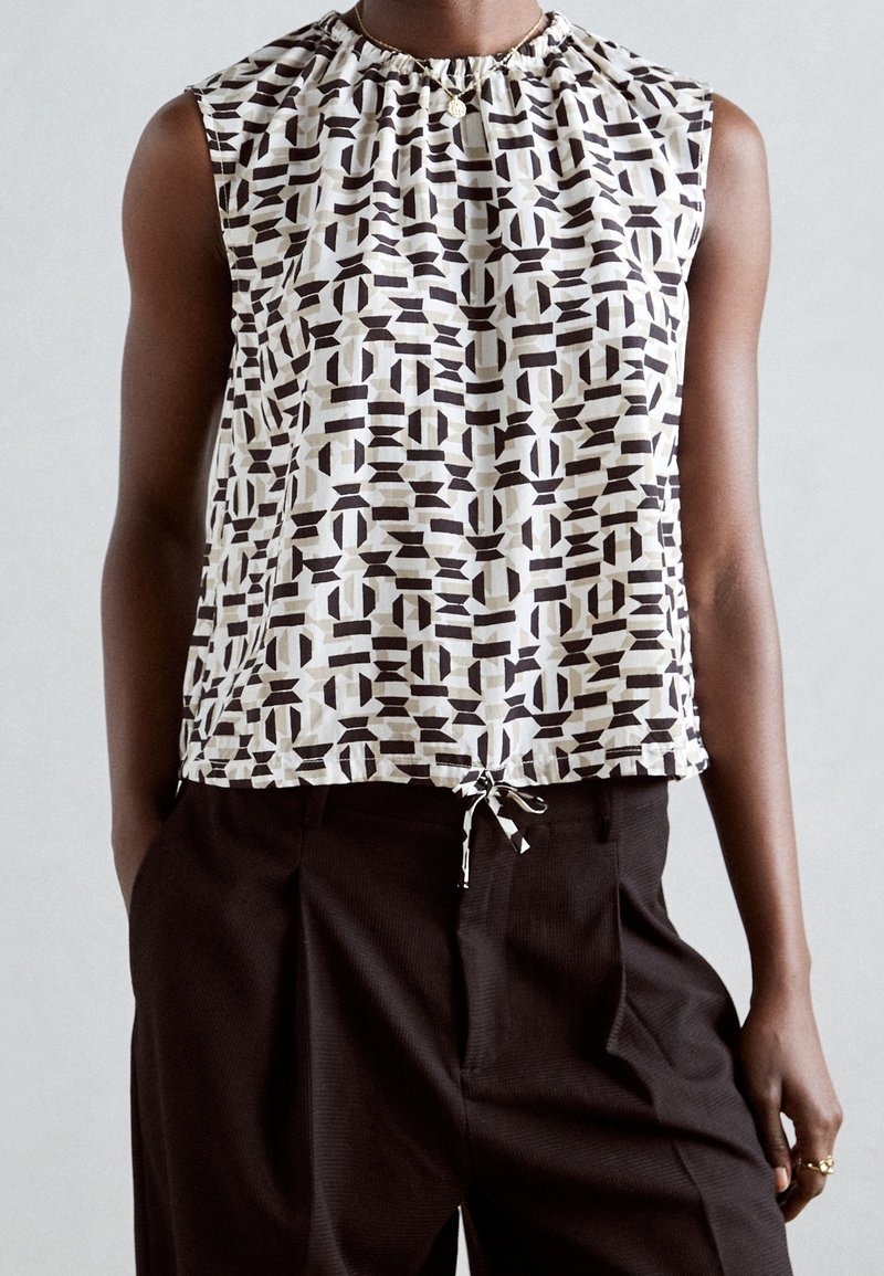 Haut sans manches avec motif géométrique noir et blanc et ourlet à cordon, porté avec un pantalon noir plissé taille haute.