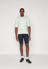T-shirt de sport vert clair à manches courtes avec une fermeture éclair à l'avant, associé à un short de cyclisme noir. Le modèle porte des chaussures noires et des chaussettes blanches.
