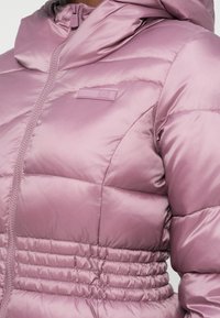 Pielāgota, pūkaina jaka maiga rozā krāsā ar rāvējslēdzēju, augstu apkakli un savilktu vidukļa detaļu. Gluda, spīdīga auduma tekstūra.